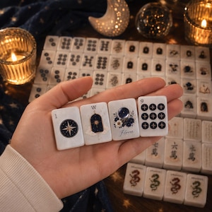 Può includere: Un set di tessere rettangolari bianche da mahjong con illustrazioni celesti e floreali. Quattro tessere sono tenute in mano, mostrando una bussola, una finestra con un sole, una sfera di cristallo con la parola Flower e nove stelle.