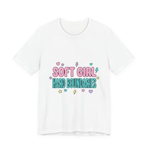 Puede incluir: Una camiseta blanca de manga corta con el texto SOFT GIRL HARD BOUNDARIES en letras de burbuja rosas y turquesas. El diseño está rodeado de pequeños iconos coloridos que incluyen corazones rosas y morados, rayos amarillos y estrellas.
