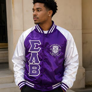 May include: A purple and white satin varsity jacket featuring the Greek letters Sigma Lambda Beta in white on the right side and a circular crest with the text Opportunity For Wisdom 1986 Wisdom For Culture on the left side with snap buttons.