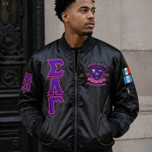 May include: A black bomber jacket featuring purple and pink Greek letters on the right chest, the text FA 20 on the right sleeve, and a crest with the text Culture Is Pride 1990 Sigma Lambda Gamma on the left chest, plus a flag patch on the left sleeve.