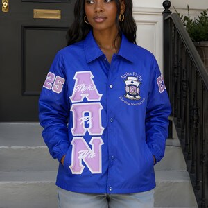 May include: A royal blue coach jacket featuring large lavender and white Greek letters on the front, the number 25 on the right sleeve, and an embroidered Alpha Pi Nu Nursing Sorority Inc. emblem on the left chest with a snap button closure.