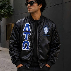 May include: A black bomber jacket featuring the Greek letters Alpha Tau Omega in blue and white on the right chest. The left chest displays a diamond-shaped emblem with intricate blue and white embroidery. The jacket has a front zipper closure.