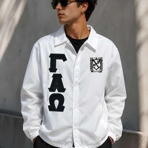May include: A white coach jacket featuring a black and green vertical letter applique on the right side and a small crest emblem on the left chest. The jacket has a classic collar, snap button closure, and elastic cuffs with a drawstring hem.