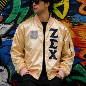 May include: A shiny gold satin bomber jacket featuring a pink ribbed collar and cuffs, a black zip, and a vertical appliqué of the Greek letters Zeta Sigma Chi in black and white. A small grey embroidered patch with a book icon is on the chest.