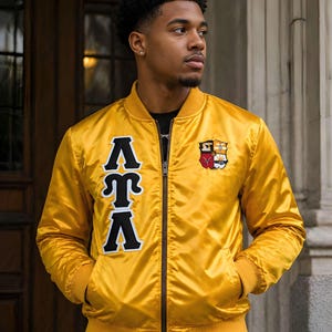 May include: A bright yellow satin bomber jacket featuring a black zipper closure, ribbed cuffs, and a ribbed hem. The right chest displays large black Greek letters with white trim, while the left chest features a colorful embroidered crest emblem.