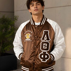 May include: A brown and white satin varsity jacket featuring the Greek letters Lambda Theta Phi in white and brown embroidery on the right side, and a Lambda Theta Phi crest with the text 1975 and Latin Fraternity Inc. on the left side.