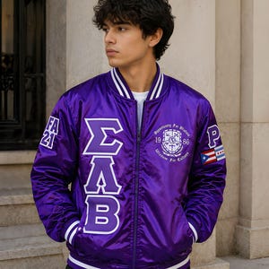 May include: A purple satin bomber jacket featuring white striped ribbed trim on the collar, cuffs, and hem. The front displays large white Greek letters, a circular crest with the text Opportunity For Wisdom 1986 Wisdom For Culture, and patches.