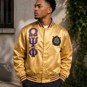 Könnte beinhalten: Eine goldene Satin-Collegejacke mit violetten griechischen Buchstaben auf der rechten Brust und einem kreisförmigen gestickten Wappen auf der linken Brust. Die Jacke hat eine Druckknopfleiste sowie gerippte Bündchen und Kragen mit Streifen.