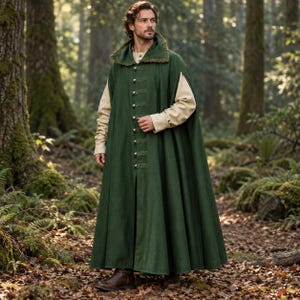 Può includere: Un mantello verde bosco lungo fino ai piedi con colletto alto dal bordo testurizzato, chiusura frontale a bottoni con linguette in tessuto orizzontali e maniche lunghe. Indossato sopra una camicia beige a maniche lunghe e stivali in pelle marrone.