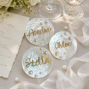 May include: Three circular white iridescent shell place cards featuring gold wire script names reading Amelia, Chloe, and Stella. Each disc is decorated with scattered white pearls of varying sizes, creating a textured and reflective surface for table settings.