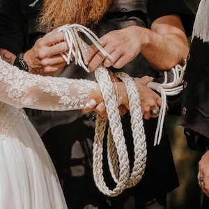 Puede incluir: Un cordón de ceremonia de mano hecho de gruesas hebras de cuerda trenzada de color crema. El cordón está construido con múltiples secciones tejidas que cuelgan en bucles, con extremos sueltos y deshilachados visibles en la parte superior.