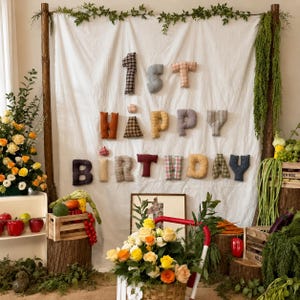 May include: A white fabric backdrop featuring plush, stuffed fabric letters spelling out 1st HAPPY BIRTHDAY in various colours and patterns. The display is framed by wooden poles with greenery, surrounded by wooden crates, fresh produce, and floral arrangements.