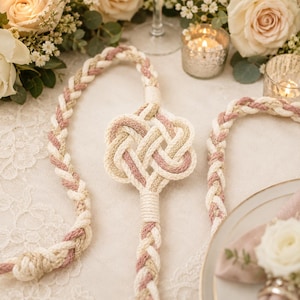 May include: A decorative rope cord featuring a complex central knot design. The cord is constructed from braided strands in cream, dusty rose, and metallic gold tones, with a thick, tightly wound binding securing the knot to the braided rope ends.