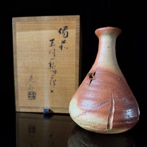 May include: A ceramic vase with a wide base and narrow neck, featuring a textured surface with mottled tan and reddish-brown tones. Behind the vase sits a rectangular wooden box with black Japanese calligraphy and a square red seal stamp.