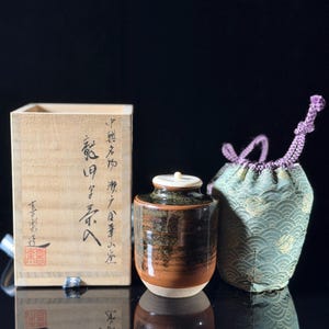 May include: A cylindrical ceramic tea caddy with a dark brown and black mottled glaze, a small white lid, a light-colored wooden storage box with black Japanese calligraphy and a red seal, and a green fabric drawstring pouch with a wave pattern.