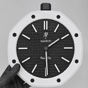 May include: An octagonal wall clock with a white bezel and a black textured dial. The face features white hour markers, white hands, and the text AP x swatch and Roy Op printed in white. The clock has a black crown at the top and eight black screws.