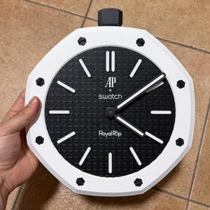 Op de afbeelding: Een achthoekige wandklok met een witte rand en een zwarte, getextureerde wijzerplaat. De wijzerplaat heeft witte uurmarkeringen, witte wijzers en de tekst AP x swatch Royal Pop. Acht zwarte schroefachtige accenten sieren de witte rand.