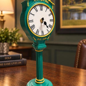 May include: A green and yellow standing clock with a white face and black Roman numerals. The top features the text TPC Piper Glen and a golf logo, while the base includes the text EST. 1988. The clock has a fluted pedestal base and black hands.