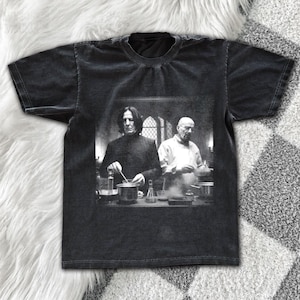 May include: A black short-sleeve t-shirt featuring a monochromatic graphic print of two figures in a laboratory setting. One figure has long dark hair and wears a dark coat, while the other is bald and wears a light-colored shirt, both working with glassware.