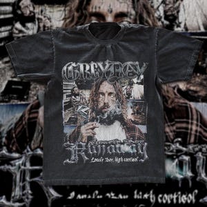 May include: A black short-sleeve t-shirt featuring a graphic print with the text GREYDAY and RUNAWAY. The design includes a central portrait of a person with long hair, a plaid shirt, and smoke, with the text Lonely Boy, high cortisol below.