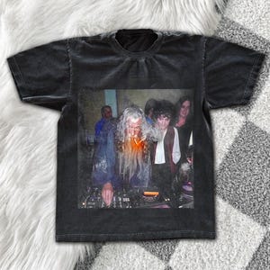 Könnte beinhalten: Ein schwarzes Kurzarm-T-Shirt mit einem Grafikdruck einer langhaarigen, bärtigen Figur, die einem Zauberer ähnelt und an einem DJ-Mischpult steht, wobei weitere Personen im Hintergrund zu sehen sind. Das T-Shirt hat eine verwaschene Optik.
