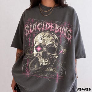 Puede incluir: Una camiseta gris carbón de manga corta con un gran estampado gráfico de un cráneo humano con un ojo rosa brillante, rodeado de serpientes. El texto "SUICIDEBOYS" aparece en una fuente rosa dentada sobre el cráneo, con "G59" impreso en la frente.