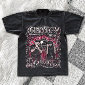 Op de afbeelding: Een zwart t-shirt met korte mouwen en een grafische print met de tekst GREYDAY NORTH AMERICA TOUR 2023. Het ontwerp bevat twee figuren, een schedel, kettingen en prikkeldraad in roze, witte en zwarte tinten op de borst.