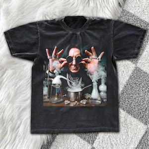 May include: A black short-sleeve t-shirt featuring a graphic print of a person with dark hair wearing round sunglasses and multiple rings. The person is surrounded by laboratory glassware, including beakers and a metal pot emitting white smoke.