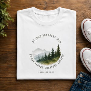 May include: A white t-shirt featuring a circular graphic with a watercolour-style mountain and forest landscape. The text "AS IRON SHARPENS IRON SO ONE PERSON SHARPENS ANOTHER" is printed in a circle around the image, with "PROVERBS 27:17" below.