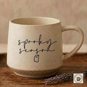 May include: A speckled cream-colored ceramic mug with a handle, featuring the black text "spooky season" and a small ghost illustration. The mug has a smooth, rounded shape with a slightly darker, solid-colored base and a speckled texture.