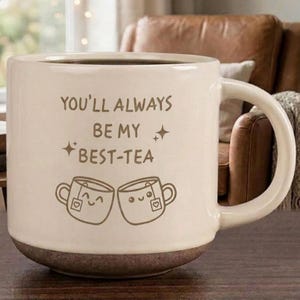 May include: A cream-colored ceramic mug with a brown base and a handle on the right side. The front features brown text that reads "YOU'LL ALWAYS BE MY BEST-TEA" with two small star icons and a graphic of two smiling teacups clinking together.