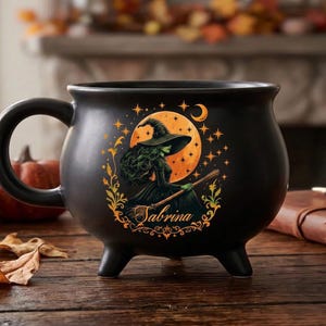 Peut inclure: Un mug noir mat en forme de chaudron orné d'une illustration représentant une sorcière à la peau verte, aux longs cheveux et au chapeau pointu, chevauchant un balai devant une grande lune orange. Le prénom Sabrina est écrit en cursive.
