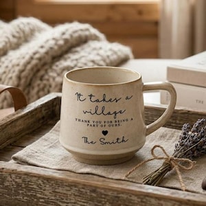 May include: A speckled cream-coloured ceramic mug with a handle, featuring the black printed text 'It takes a village, thank you for being a part of ours, The Smith' and a small black heart icon centred on the front of the vessel.