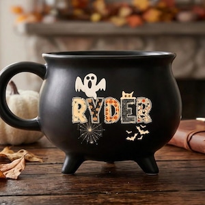 May include: A matte black cauldron-shaped mug featuring the name RYDER in patterned block letters. The design includes a white ghost, a spider web, a cat peeking over the letters, and small bats. The mug has three short legs and a curved handle.