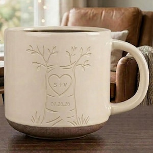 May include: A cream-coloured ceramic mug with a dark brown base, featuring an embossed tree design on the front. The tree trunk displays a heart shape with the letters S + V inside and the date 07.26.26 engraved below the heart symbol.