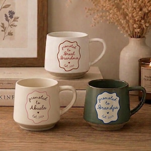 May include: Three ceramic mugs with handles. Two are off-white and one is sage green. Each features a wavy-edged frame containing text: Promoted to Great Grandma Est. 2027 in red, Promoted to Abuela Est. 2026 in pink, and Promoted to Grandpa Est. 2027 in blue.