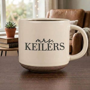May include: A cream-coloured ceramic mug with a dark brown base and a handle on the right side. The front of the mug features the black text "mrs. KEILERS" printed in a combination of script and serif fonts, centred on the smooth surface.