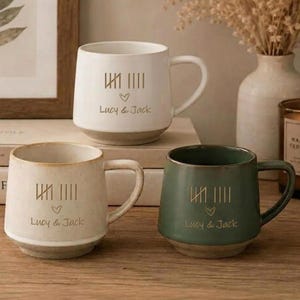 May include: Three ceramic mugs in white, cream, and sage green, each featuring a gold-colored tally mark design, a small heart icon, and the text "Lucy & Jack" printed on the front. The mugs have a smooth finish and a sturdy handle on the side.