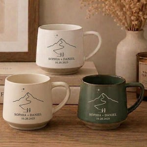 May include: Three ceramic mugs with handles, two in off-white and one in sage green. Each mug features a black line drawing of a mountain range with a path and two figures, plus the printed text SOPHIA + DANIEL and the date 10.20.2025.