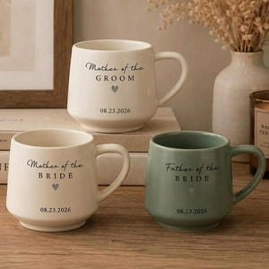 May include: Three ceramic mugs with handles, two in cream and one in sage green. Each mug features black text reading Mother of the Groom, Mother of the Bride, or Father of the Bride, a small heart icon, and the date 08.23.2026 printed on the front.