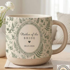 May include: A ceramic mug with a speckled cream base and a green botanical fern and floral pattern. The center features an oval frame containing the text Mother of the BRIDE, a small heart icon, and the date 08.23.2026 in dark grey lettering.
