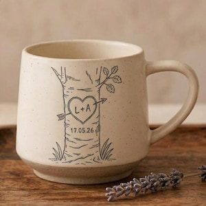 May include: A cream-colored ceramic mug with a speckled finish and a single handle. The front features a black line drawing of a tree trunk with a heart carved into the bark containing the letters L+A and an arrow, with the date 17.05.26 below.