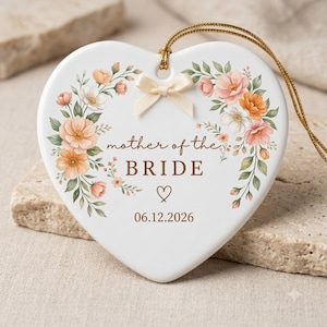 May include: A white heart-shaped ceramic ornament featuring a floral wreath of peach, orange, and white flowers. The text reads "mother of the BRIDE" above a small heart icon and the date "06.12.2026". A cream ribbon bow and gold hanging cord are attached.
