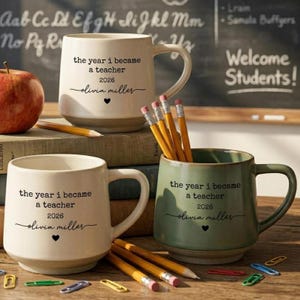 May include: Two ceramic mugs, one cream and one sage green, each printed with the text 'the year i became a teacher 2026' and 'olivia miller' with a heart icon. Several yellow pencils and colorful plastic paper clips are scattered nearby.