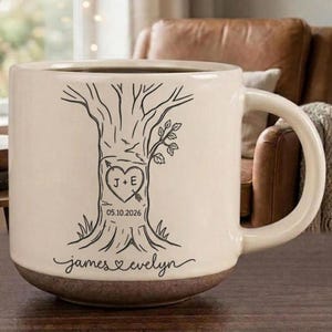 May include: A cream-colored ceramic mug with a dark brown base, featuring a black line drawing of a tree trunk. A heart carved into the tree contains the letters J + E and the date 05.10.2026. The names James and Evelyn are written below.
