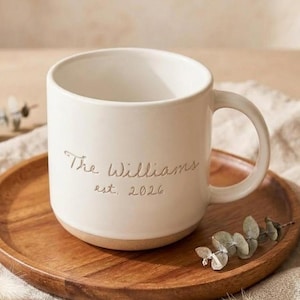 May include: A cream-colored ceramic mug with a rounded handle and a smooth finish. The front of the mug features the engraved text "The Williams" above the text "est. 2026" in a simple, clean font. The base of the mug is unglazed and tan.