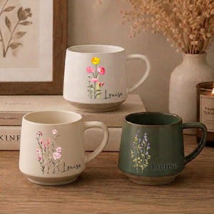 May include: Three ceramic mugs with handles, each featuring a unique wildflower illustration and the name Louise printed in script. One mug is white with pink and yellow flowers, one is cream with pink flowers, and one is dark green with purple flowers.
