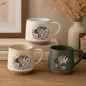 May include: Three ceramic mugs featuring a raccoon illustration and the name "jimothy" printed above the animal. One mug is white, one is cream, and one is sage green. Each mug has a handle and a floral wreath design surrounding the raccoon.