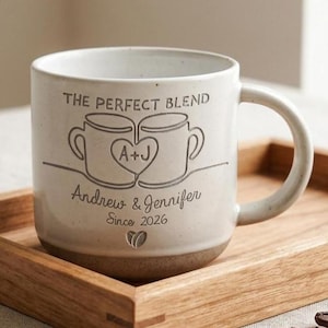 May include: A speckled off-white ceramic mug featuring the engraved text THE PERFECT BLEND, a graphic of two mugs joined by a heart with A+J inside, the names Andrew & Jennifer, the text Since 2026, and a small coffee bean icon at the bottom.