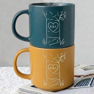 May include: A stack of two ceramic mugs, one teal and one mustard yellow, each featuring a white line-art illustration of a tree trunk with a heart carved into the centre containing the letters A & C in a clean, simple sans-serif font.